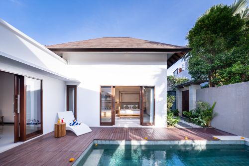Modern Balinese villa with private infinity pool, wooden deck lounge chair, open bedroom doors, and lush tropical garden under blue sky.