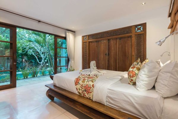 Luxurious Balinese bedroom with carved wooden bed, floral bedding, swan towel art, and tropical garden view through open sliding doors.