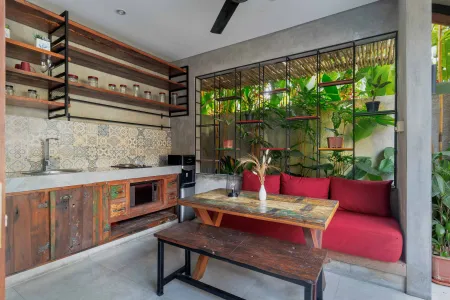 Rustic tropical kitchen with wooden cabinets, shelves of jars, tiled sink, and lush green plant wall behind metal frame, plus wooden dining bench and table. - Bali Villas