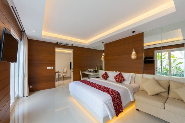 Spacious modern Balinese-style hotel bedroom with king bed, wooden walls, beige sofa, and garden-view balcony.