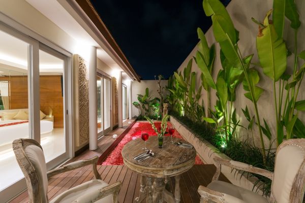 Luxurious Balinese villa terrace at night: wooden table with red petals, white armchairs, tropical banana plants, and sliding glass doors to bedroom. - Bali Villas