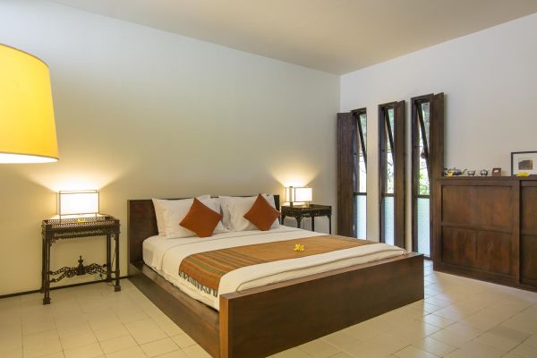 Serene Balinese bedroom interior with wooden platform bed, orange pillows, woven throw, and warm lamp lighting. - Bali Villas