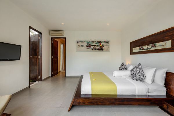 Spacious Balinese villa bedroom with low wooden platform bed, yellow runner, plush pillows, wall-mounted TV, and abstract artwork.