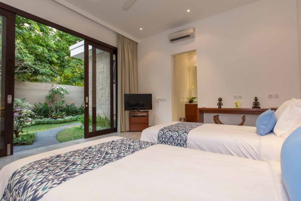 Spacious tropical bedroom with twin beds, blue bedding, flat-screen TV, and sliding doors opening to a lush garden.