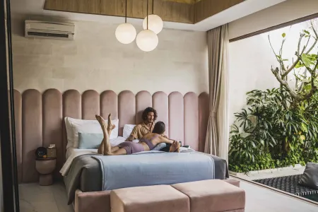 A couple in underwear cuddling intimately on a pink-tufted bed in a luxurious modern bedroom with garden views and pendant lights.