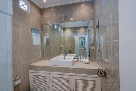 Modern bathroom interior with beige tiled walls, large frameless mirror, white vanity sink, toiletries, and walk-in shower. - Bali Villas