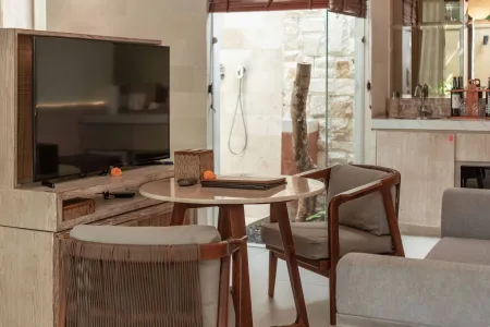 Modern tropical villa interior with wooden TV cabinet, round table with oranges, rattan chairs, gray sofa, and open stone-walled bathroom. - Bali Villas