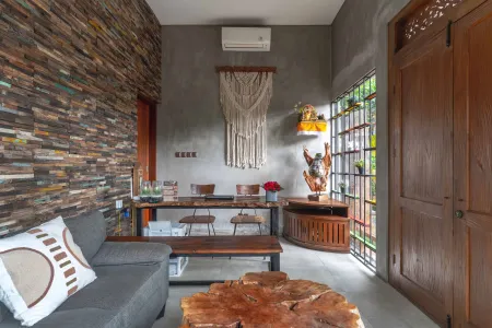 Modern tropical living room with reclaimed wood accent wall, gray sofa, tree stump coffee table, wooden dining set, macramé hanging, and potted plants. - Bali Villas