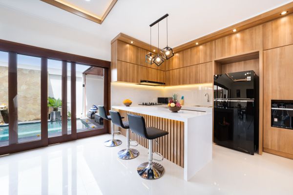 Modern wooden kitchen with white island, pendant lights, black fridge, and pool view through glass doors. - Bali Villas