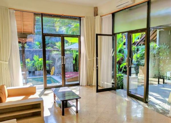 Bright Balinese villa interior with sheer curtains, beige sofa, and large glass doors opening to a lush tropical garden and plunge pool. - Bali Villas