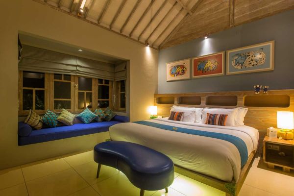 Cozy Balinese villa bedroom featuring a king-sized bed with colorful accents, blue cushioned window seat, abstract art, and warm wooden accents.