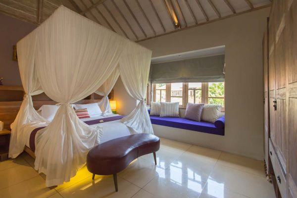 Luxurious Balinese bedroom with white canopy four-poster bed, wooden accents, purple window seat, and tropical window view.
