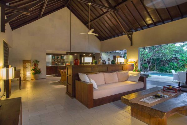 Spacious Balinese villa living room with wooden accents, beige sofa, open kitchen, and views of tropical pool garden. - Bali Villas