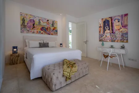 Spacious modern Balinese-style bedroom with king bed, vibrant abstract wall art, white desk, tufted ottoman, and open door to garden.