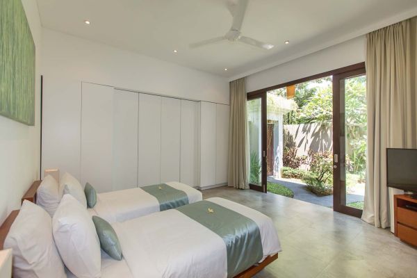 Modern Bali villa bedroom with two white twin beds, teal accents, open sliding doors to lush tropical garden, and abstract green wall art.