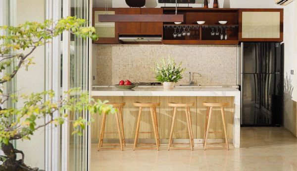 Modern wooden kitchen with island counter, bar stools, fresh flowers and apples, tiled backsplash, and open shelving in a bright, airy interior. - Bali Villas