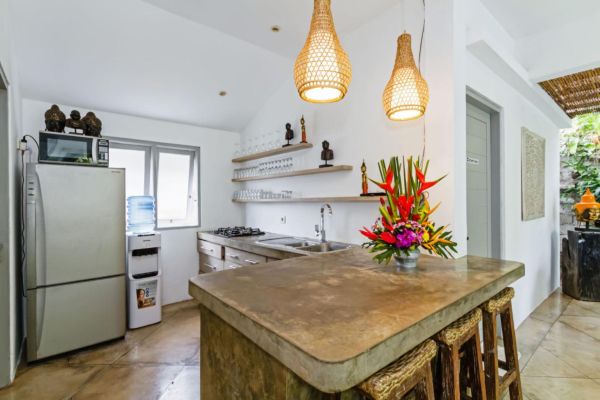 Modern Balinese-style kitchen with bamboo pendant lights, concrete island bar, white cabinets, tropical flower vase, and wooden statues. - Bali Villas