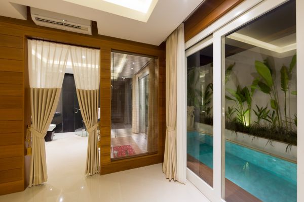 Modern wooden interior with sheer-curtained sliding doors to a bathroom and an outdoor turquoise pool amid tropical plants. - Bali Villas