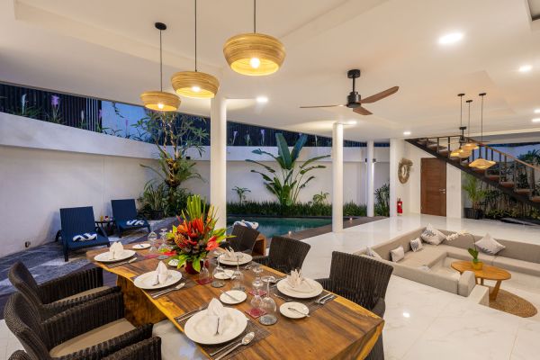 Luxurious Balinese villa interior with wooden dining table set for dinner, infinity pool, lounge chairs, and tropical plants under pendant lights at dusk. - Bali Villas