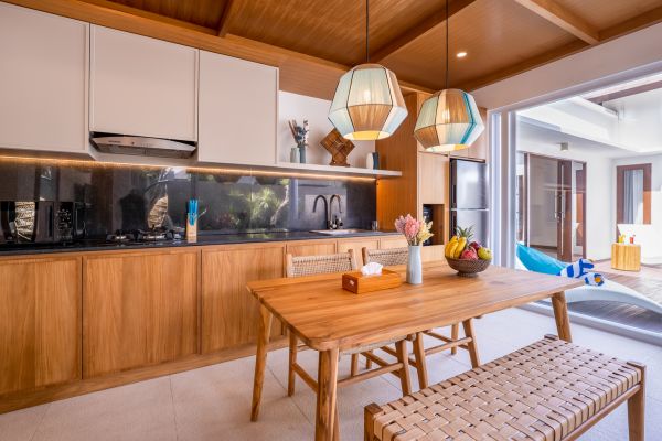 Modern tropical kitchen with wooden cabinets, pendant lights, and dining table overlooking poolside patio. - Bali Villas
