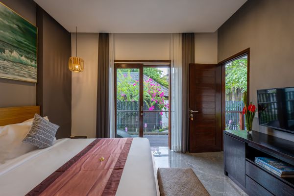 Luxurious Balinese bedroom with king-sized bed, tropical garden view through sliding glass doors, and modern decor including artwork and vase of red tulips.