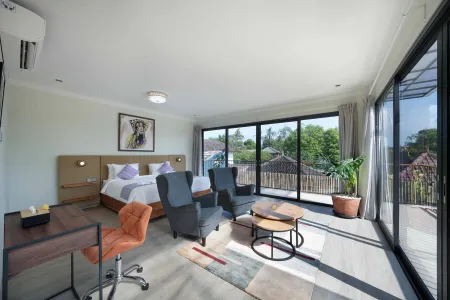 Spacious modern bedroom suite with king bed, lounge seating, desk, and sliding doors opening to a tropical balcony view.