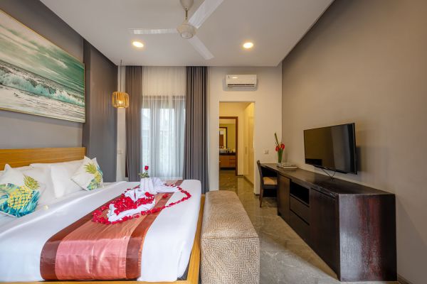 Luxurious Balinese bedroom suite with king bed topped by rose petals and red sash, seascape wall art, ceiling fan, TV, and sheer-curtained windows.