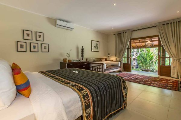Spacious Balinese bedroom with king bed, colorful pillows, wood furniture, and open doors revealing tropical garden.