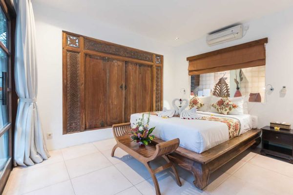 Serene Balinese bedroom with intricately carved wooden bed, white floral bedding, carved wall panels, flower-adorned bench, and sheer curtains.
