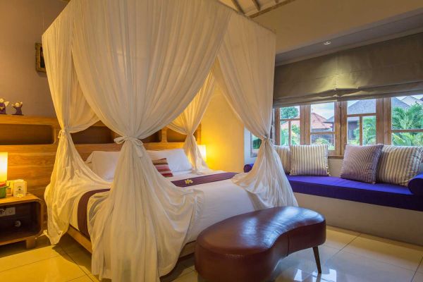 Luxurious Balinese bedroom with white canopy bed, red duvet, purple bench, and tropical window view.