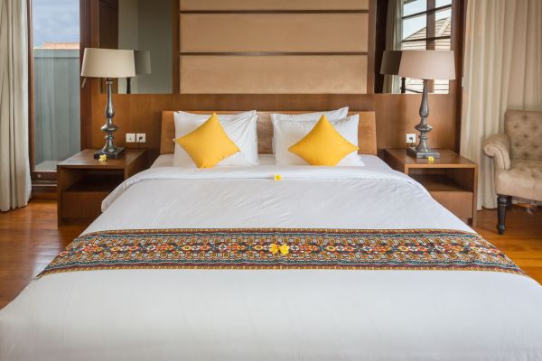 Elegant Balinese bedroom with white king bed, yellow pillows, ornate runner, and wooden accents.