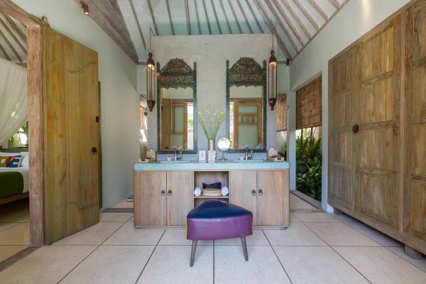 Elegant Balinese-style bathroom with wooden double vanity, carved mirrors, purple stool, and open doors to a lush bedroom.