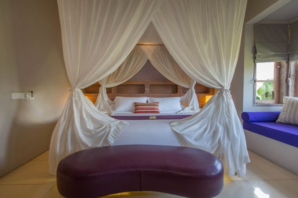 Elegant four-poster canopy bed with flowing white drapes in a serene tropical bedroom, featuring purple ottoman and blue window bench.