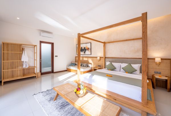 Elegant Balinese bedroom with wooden four-poster bed, green linens, tropical flower offerings, and fruit basket on a low table.