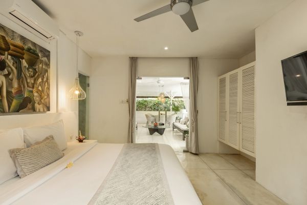 Luxurious Bali villa bedroom with white king bed, Balinese wall art, ceiling fan, and glass doors opening to tropical garden balcony.