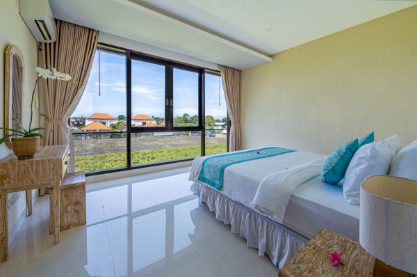 Spacious Balinese bedroom with panoramic view of green fields through floor-to-ceiling windows, king bed with teal bedding, wooden furniture, and orchids.