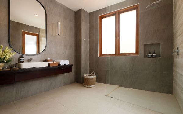 Modern gray-tiled bathroom with wooden vanity, oval black mirror, open shower, and tropical plant. - Bali Villas