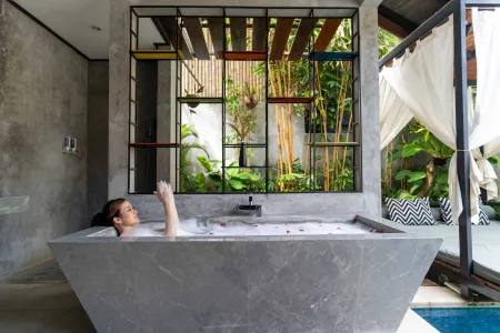 Woman relaxing in a luxurious gray stone bathtub with flower petals, overlooking a tropical garden and pool through a metal-framed window. - Bali Villas