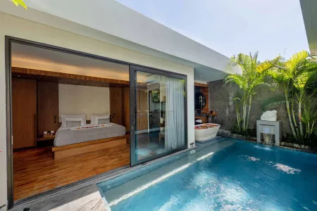 Luxurious tropical villa bedroom with king bed, open sliding doors to private plunge pool, outdoor bathtub, and lush palm greenery under blue sky.