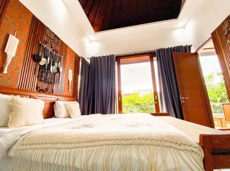 Balinese-style bedroom interior with carved wooden walls, king bed in white linens, and large glass doors opening to a tropical balcony view.