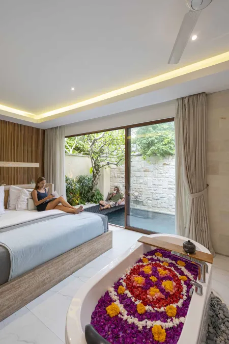 Luxurious Balinese villa bedroom with a woman reading on a bed, overlooking a lush garden, and a freestanding bathtub filled with vibrant purple and pink floating flowers.