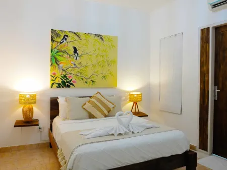 Cozy tropical bedroom with wooden king bed, swan-shaped towels, rattan lamps, and vibrant yellow bamboo bird artwork on white walls. - Bali Villas