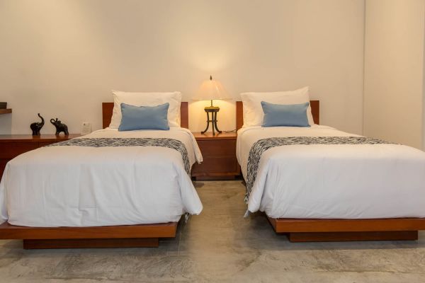 Cozy twin bedroom with wooden beds, white bedding accented in blue, bedside lamp, and elephant statues on light walls.