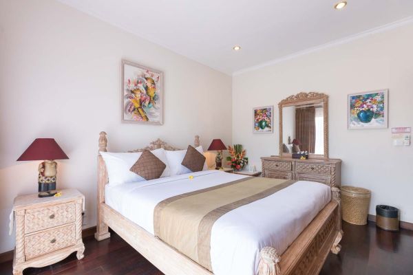 Elegant Balinese-style bedroom with carved wooden four-poster bed, white linens, bedside lamps, floral artwork, and tropical decor.