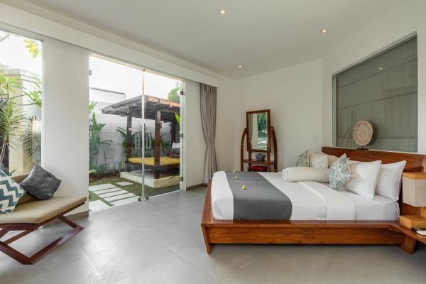 Spacious modern tropical bedroom with wooden king bed, gray linens, and open sliding doors to lush garden courtyard.