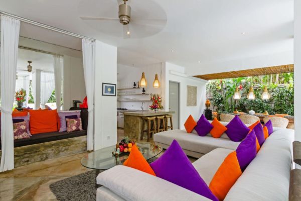 Spacious Bali-style living room with white sofa accented by orange and purple cushions, glass coffee table, open kitchen, and lush tropical garden view. - Bali Villas