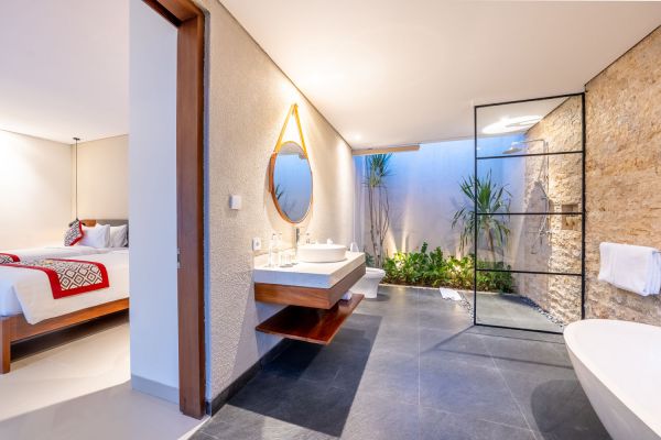 Spacious Balinese-style open-plan bathroom and bedroom suite with wooden accents, double vanity, round mirror, freestanding tub, glass shower, and tropical plants.