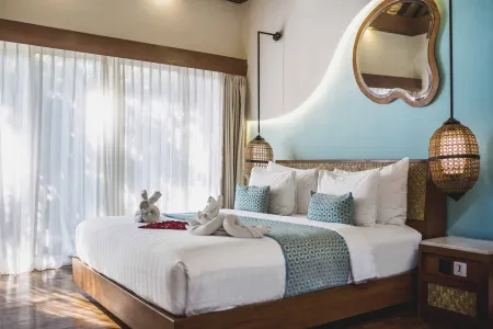 Luxurious tropical bedroom with wooden king bed, white linens, turquoise pillows, stuffed bunnies, and natural light through sheer curtains.