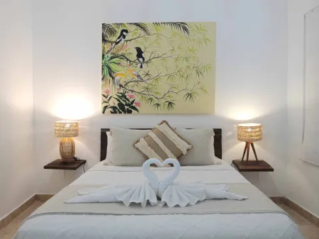 Cozy tropical bedroom featuring heart-shaped swan towel art on white bedding, flanked by rattan lamps and a vibrant painting of birds in foliage above.