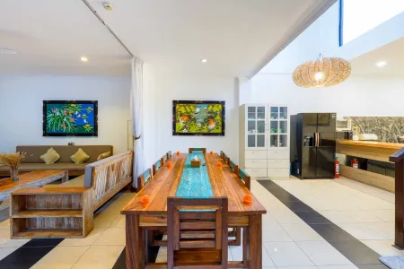 Spacious open-plan dining area with wooden table, tropical wall art, beige sofa, and modern kitchen in Balinese-style interior. - Bali Villas